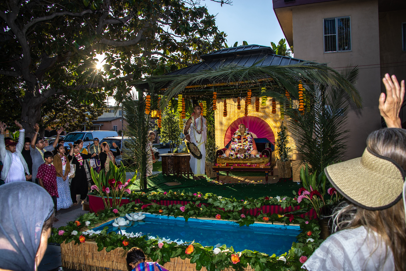 Prabhupada Festival at New Dwarka Temple -- devotees gathered around the decorated vyasasana with marigold garlands, lotus pool, and sunset light